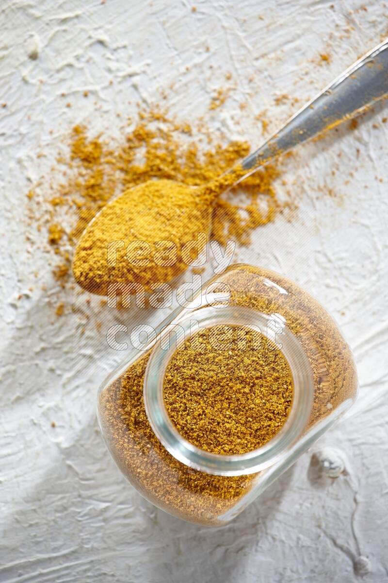 A glass spice jar and metal spoon full of turmeric powder on textured white flooring