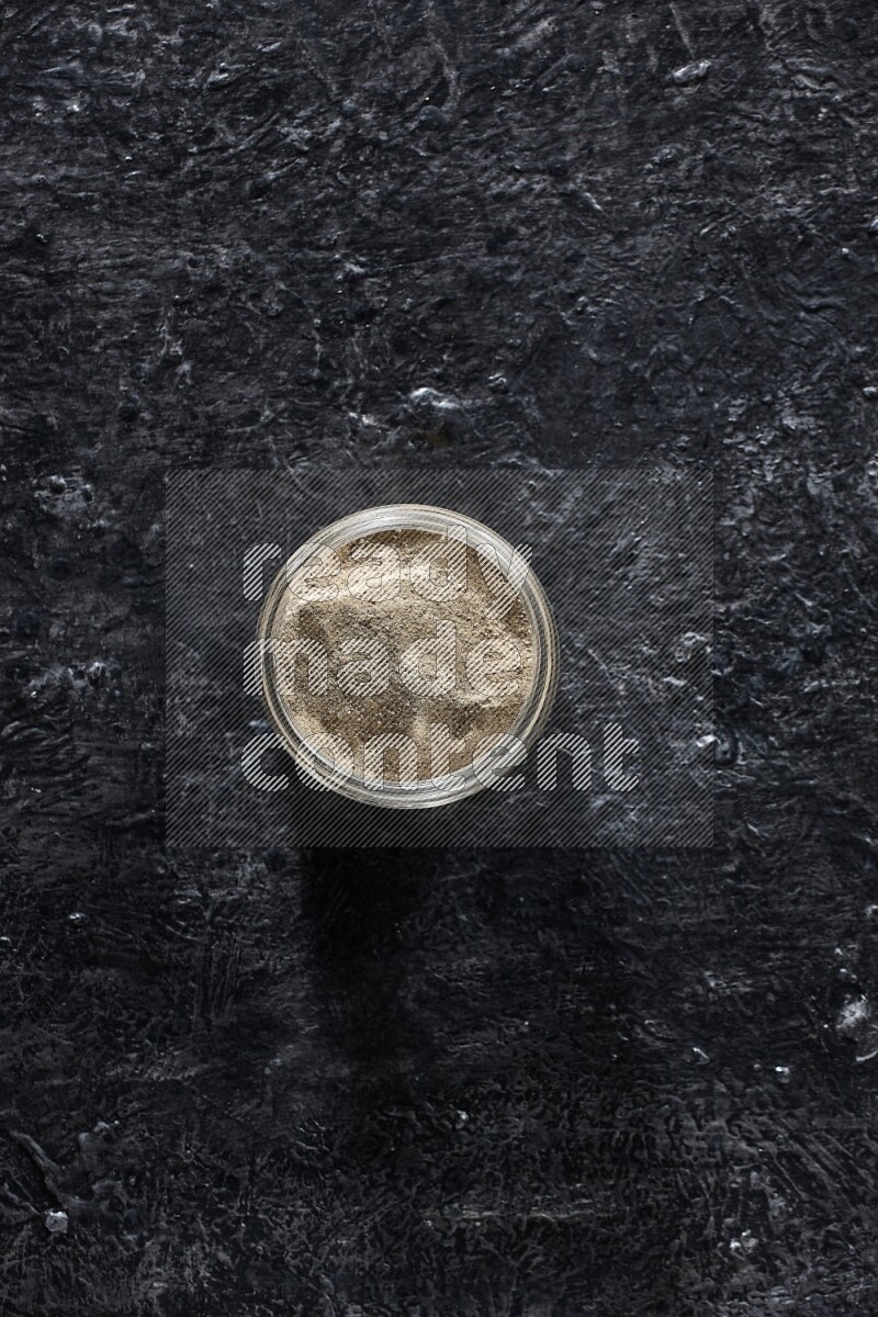A glass jar full of white pepper powder on textured black flooring