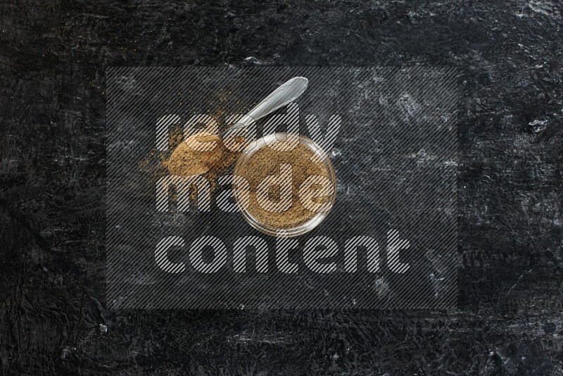 وعاء زجاجي للتوابل وملعقة معدنية ممتلئين ببودرة الكمون على خلفية سوداء