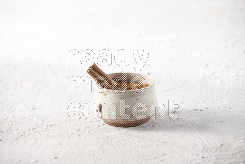 Ceramic beige bowl full of cinnamon powder with a cinnamon stick on a textured white background