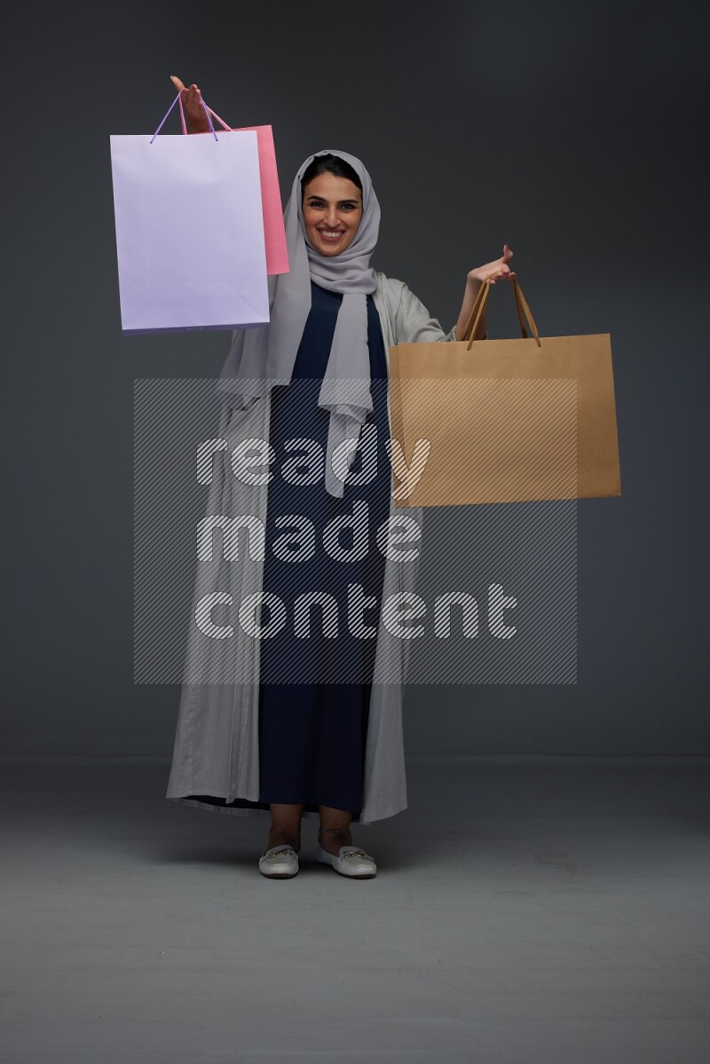 A Saudi woman wearing a light gray Abaya and head scarf standing and holding shopping bags on a grey background