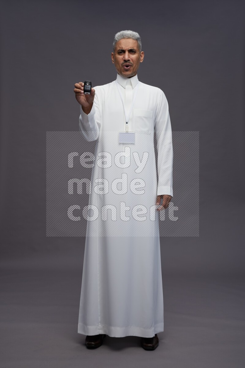 Saudi man wearing thob with neck strap employee badge standing holding key car on gray background