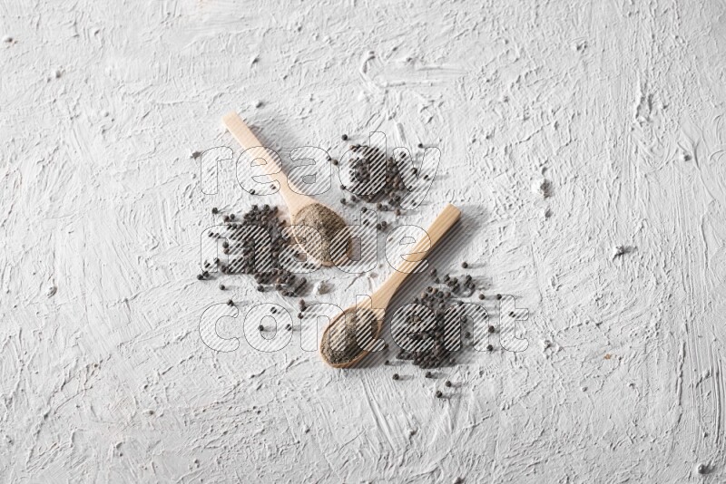 2 wooden spoons full of black pepper powder and black pepper beads spread on textured white background