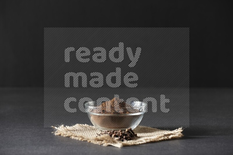 A glass bowl full of cloves powder with cloves grains on a burlap piece on a black flooring