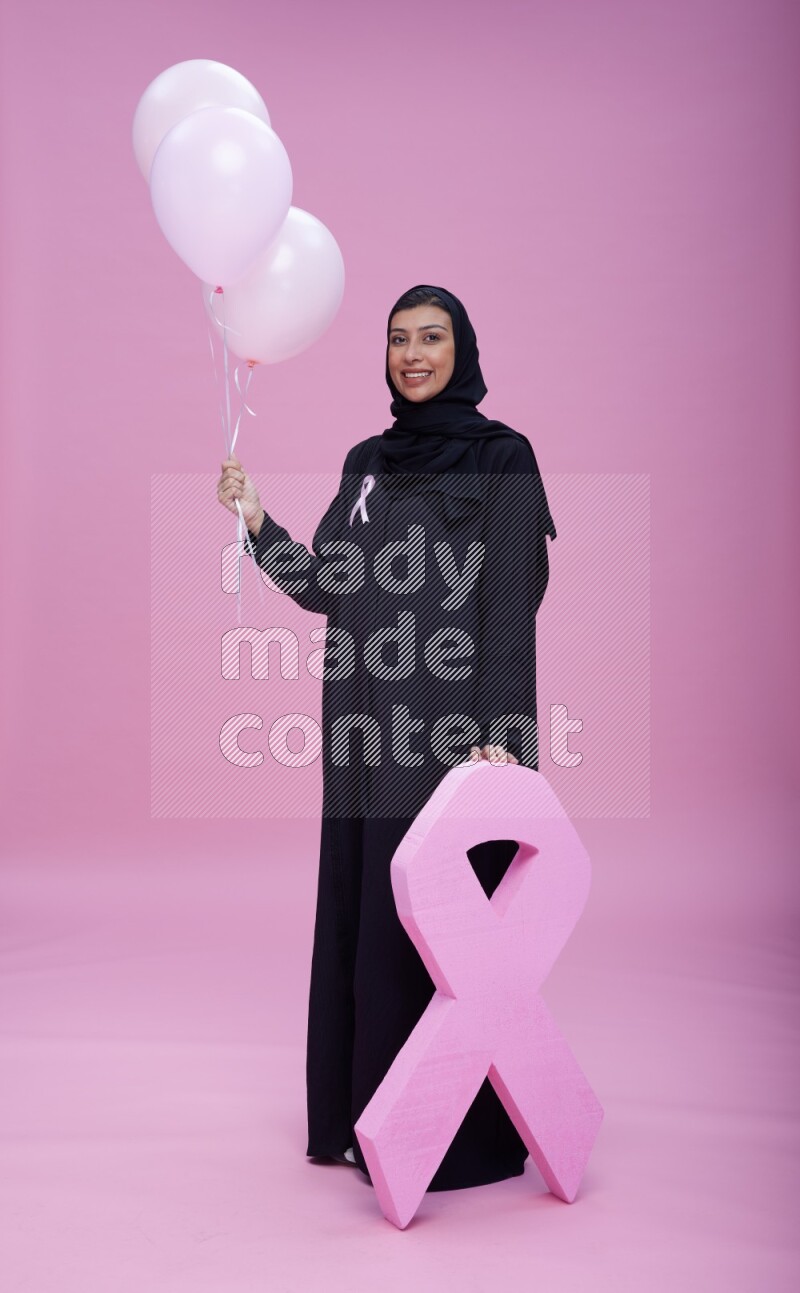 Saudi woman wearing pink ribbon on Abaya standing holding balloons and awareness sign on pink background