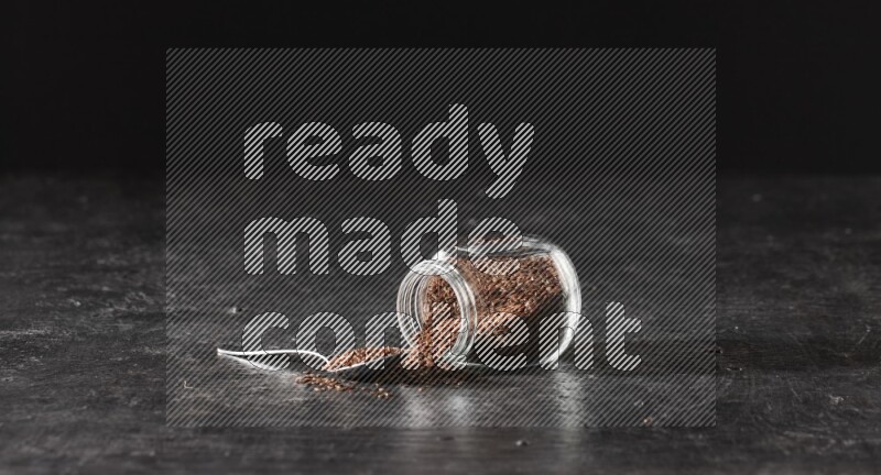 A glass spice jar full of flaxseeds flipped and seeds spread out with a metal spoon full of the seeds on a textured black flooring