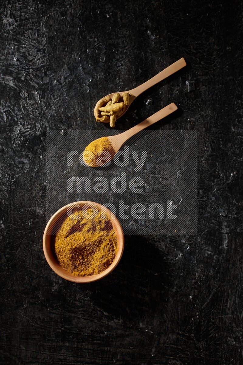 A wooden bowl full of turmeric powder and 2 wooden spoons full of dried turmeric finger and turmeric powder on textured black flooring