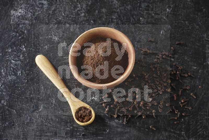 A wooden bowl and wooden spoon full of cloves powder with spreaded cloves on textured black flooring