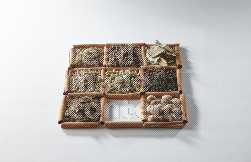 9 squares of cinnamon sticks full of cardamom in the middle surrounded by nutmeg, salt, dried basil, white pepper, cumin, allspice, bay laurel leaves and black pepper on white flooring