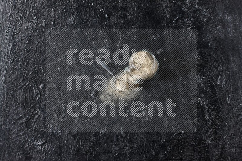 وعاء زجاجي عشبي مقلوب وملعقة معدنية ممتلئة ببودرة الفلفل الأبيض مع بودرة متناثرة على أرضية سوداء