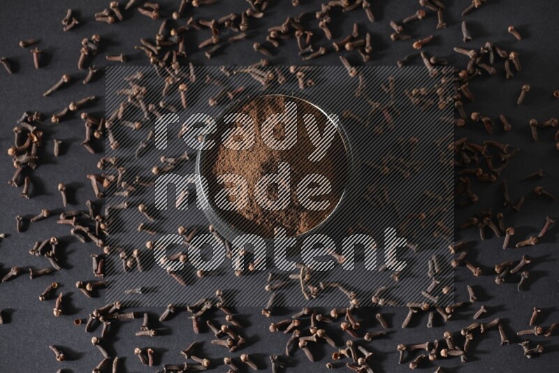 A black pottery bowl full of cloves powder and cloves whole grains on a black flooring