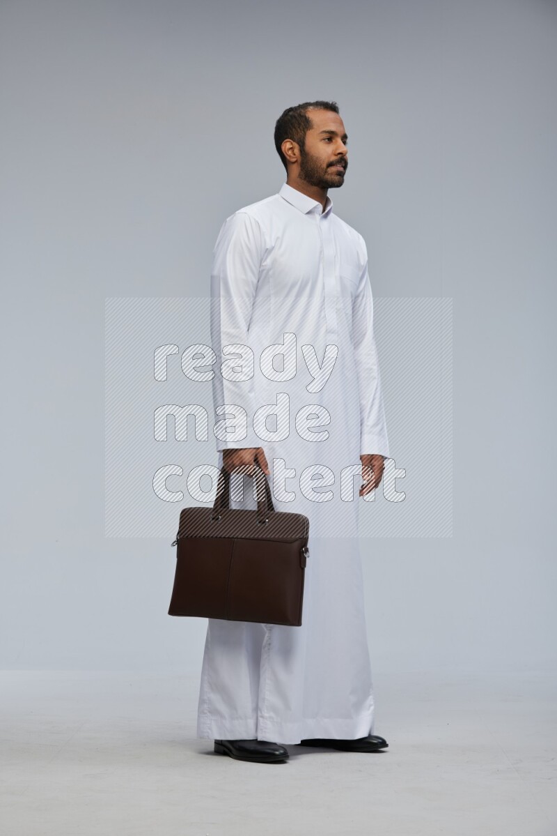 Saudi man wearing thob standing holding suitcase on gray background