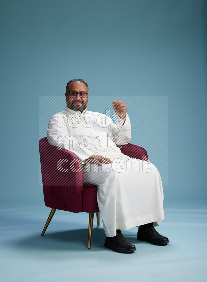 Saudi Man without shimag sitting on chair Interacting with the camera on blue background