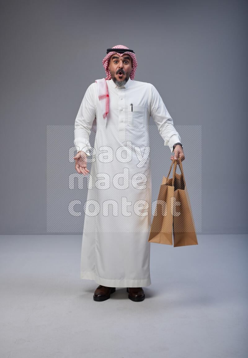 Saudi man Wearing Thob and red Shomag standing holding shopping bag on Gray background