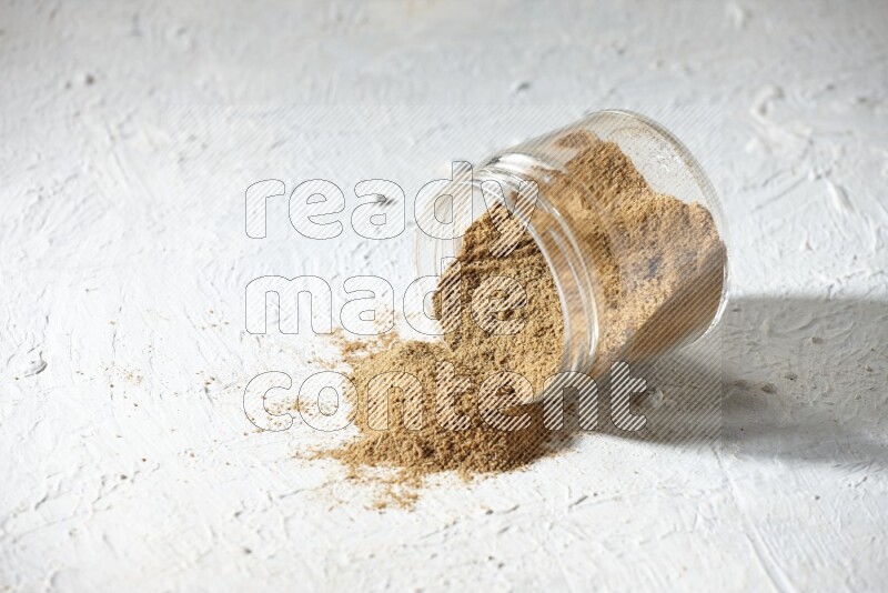 A flipped glass jar full of cumin powder and powder spilled out on textured white flooring