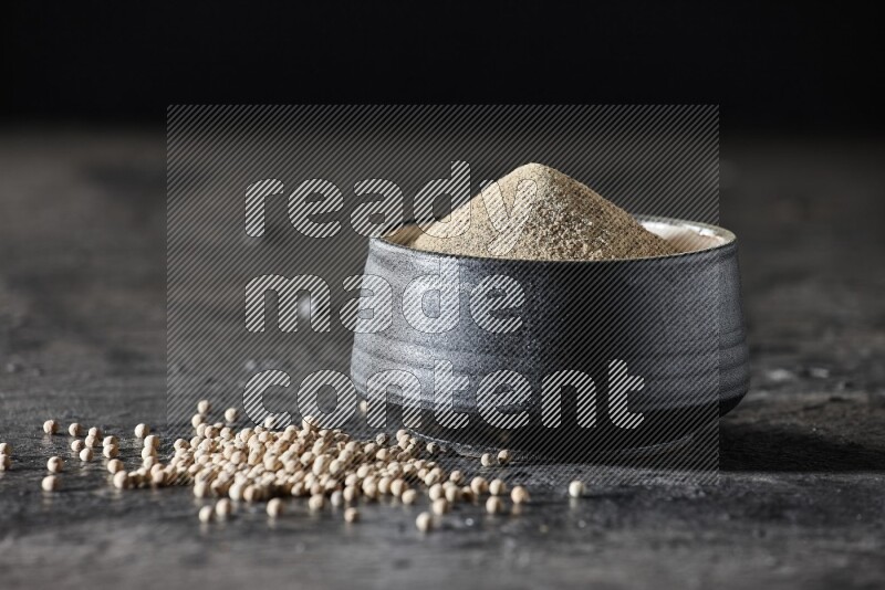 Black pottery bowl full of white pepper powder with white pepper beads on textured black flooring