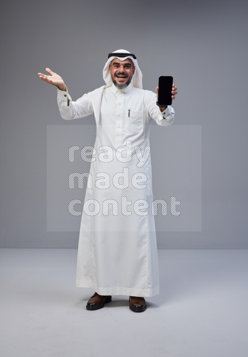 Saudi man Wearing Thob and white Shomag standing showing phone to camera on Gray background