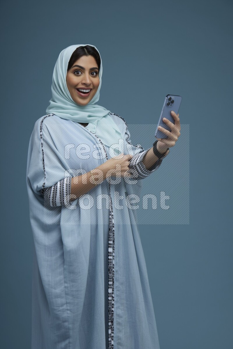 A Saudi woman posing with her phone in a blue background wearing a blue Abaya with hijab