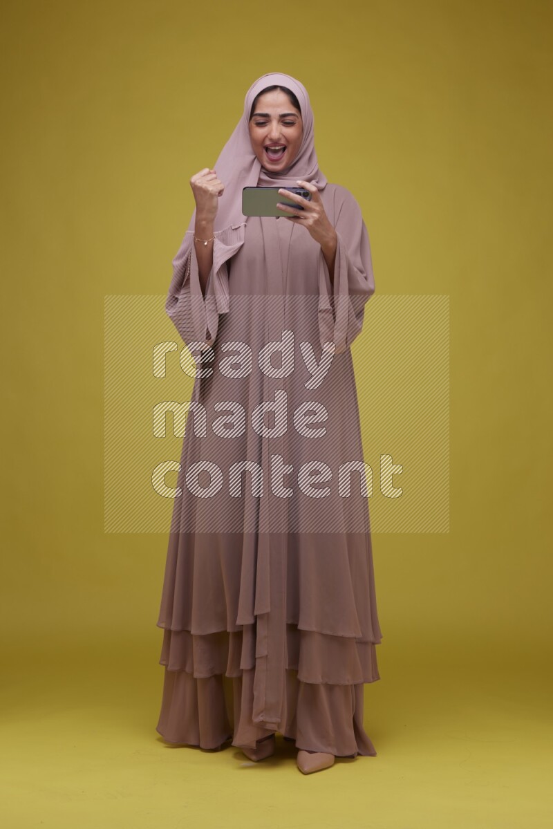 A woman Playing Games a Yellow Background wearing Brown Abaya with Hijab