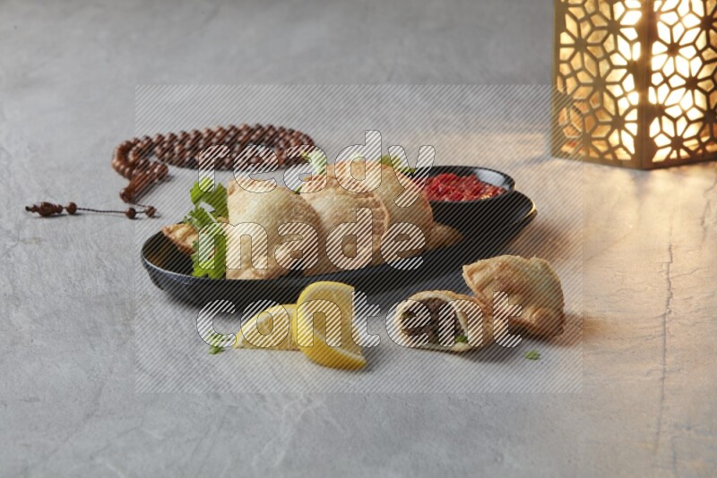 Five fried sambosas in an oval shaped black plate, beside a cut meat sambosa, a brown misbaha and a golden lantern on a gray background