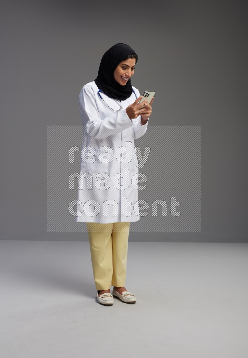Saudi woman wearing lab coat with stethoscope standing texting on phone on Gray background