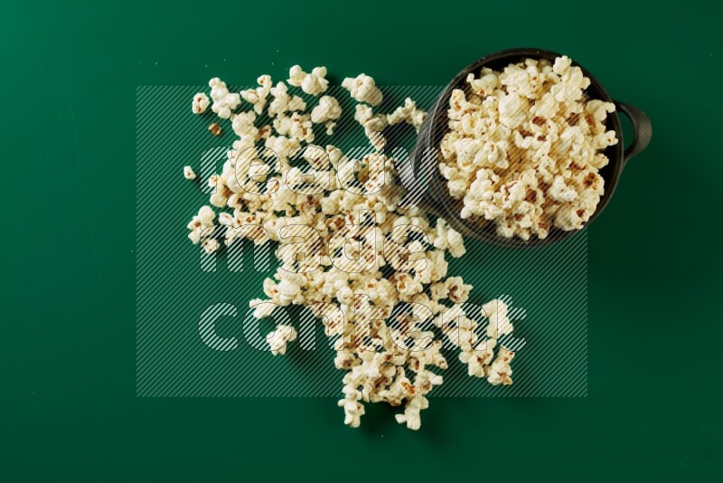 A black ceramic bowl full of popcorn with popcorn beside it on a green background in a top view shot