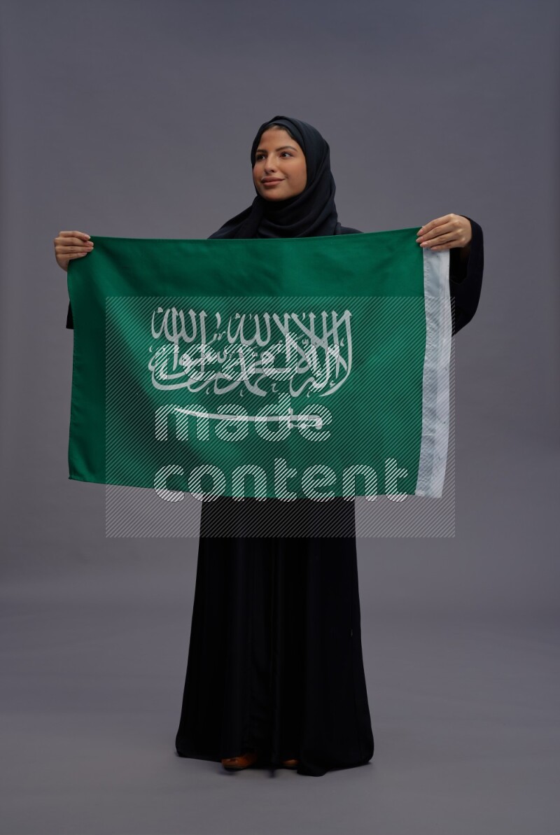 Saudi woman wearing Abaya standing holding Saudi flag on gray background