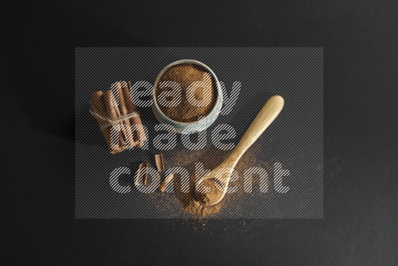 Ceramic bowl full of cinnamon powder and a wooden spoon full of powder with cinnamon sticks stacked and bounded on black background