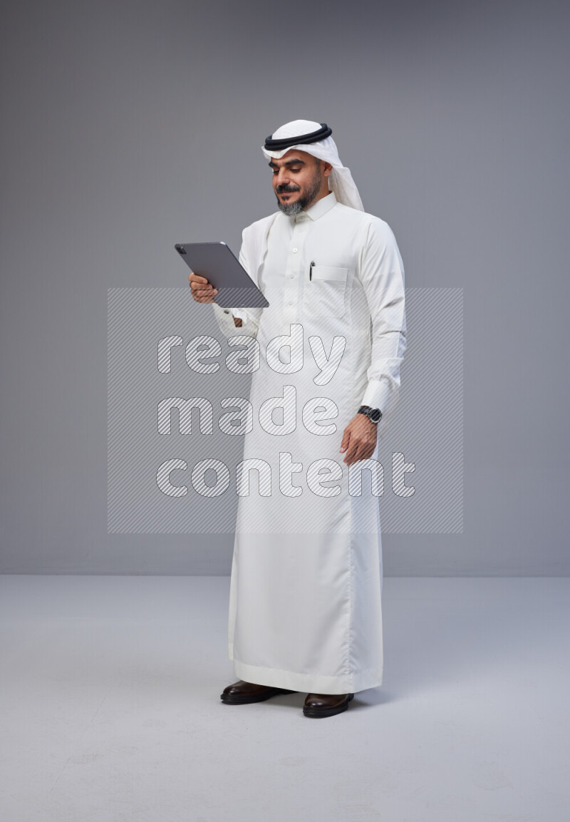 Saudi man Wearing Thob and white Shomag standing working on tablet on Gray background