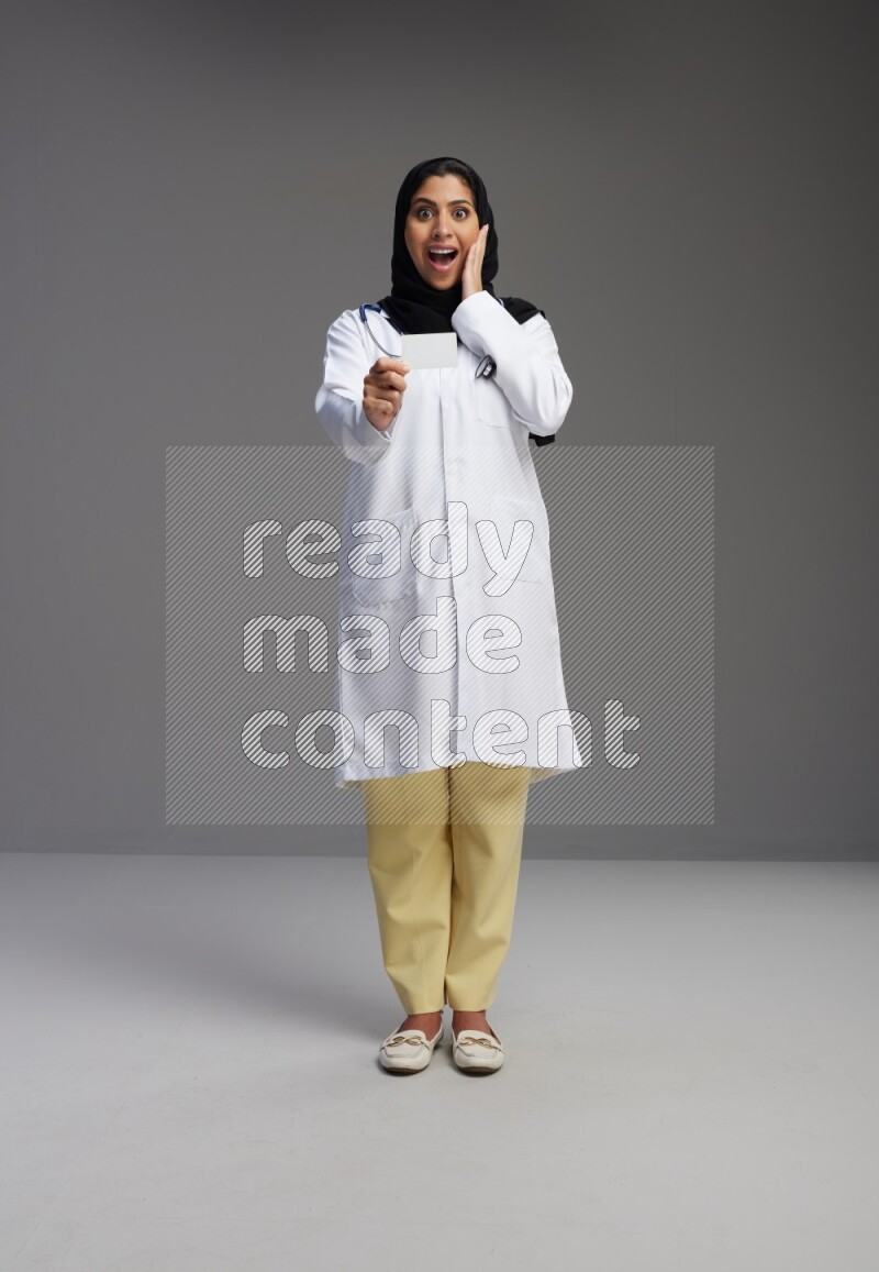 Saudi woman wearing lab coat with stethoscope standing holding ATM card on Gray background