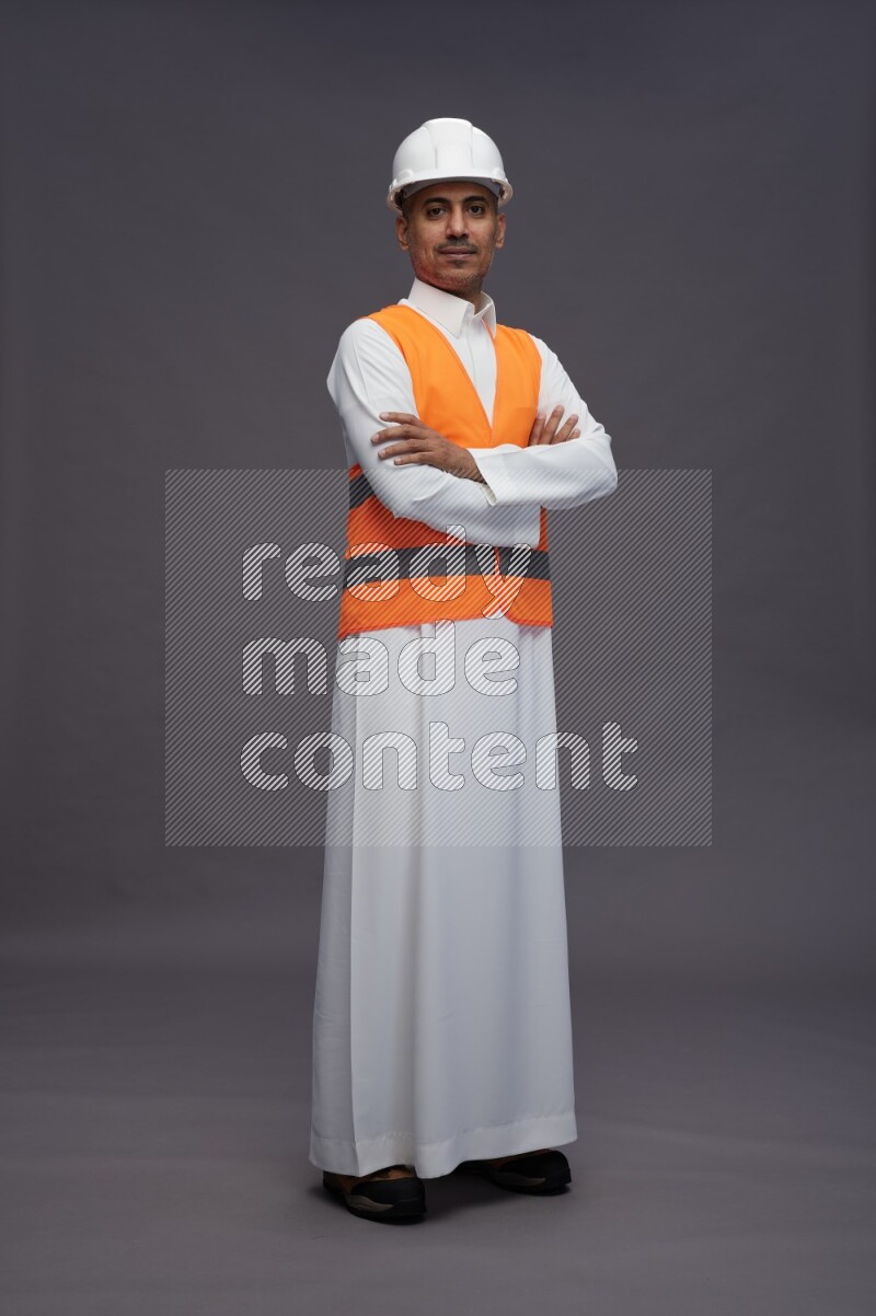 Saudi man wearing thob with engineer vest standing with crossed arms on gray background