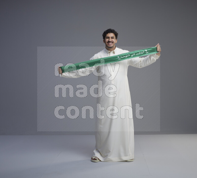 A Saudi man standing wearing thob holding Saudi flag scarf on gray background