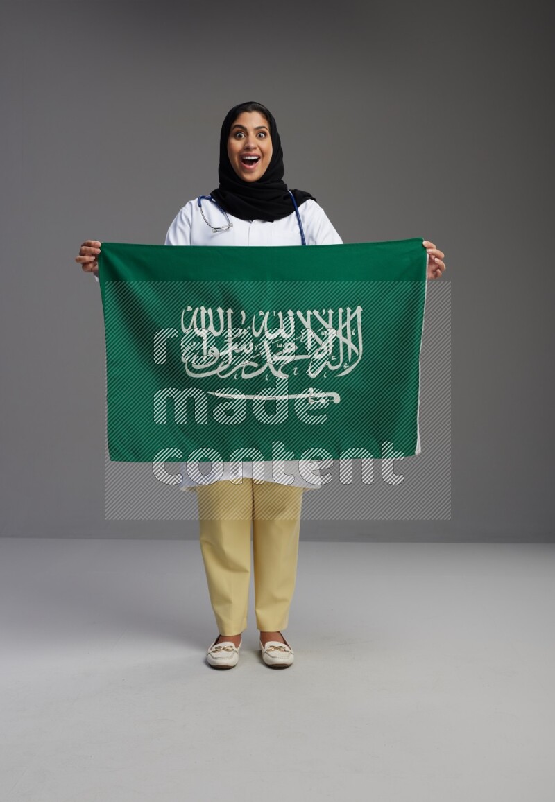 Saudi woman wearing lab coat with stethoscope standing holding Saudi flag on Gray background