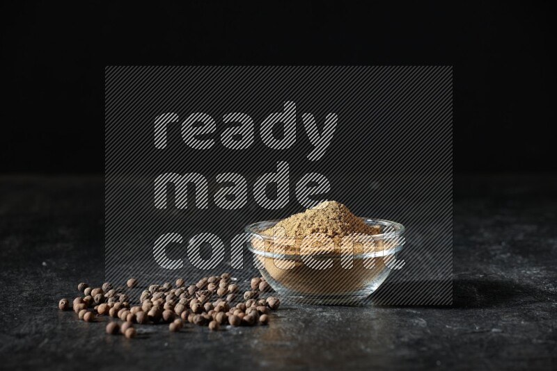 وعاء زجاجي ممتلئ ببودرة بهار الفلفل الحلو وحبوبه متناثرة على خلفية سوداء