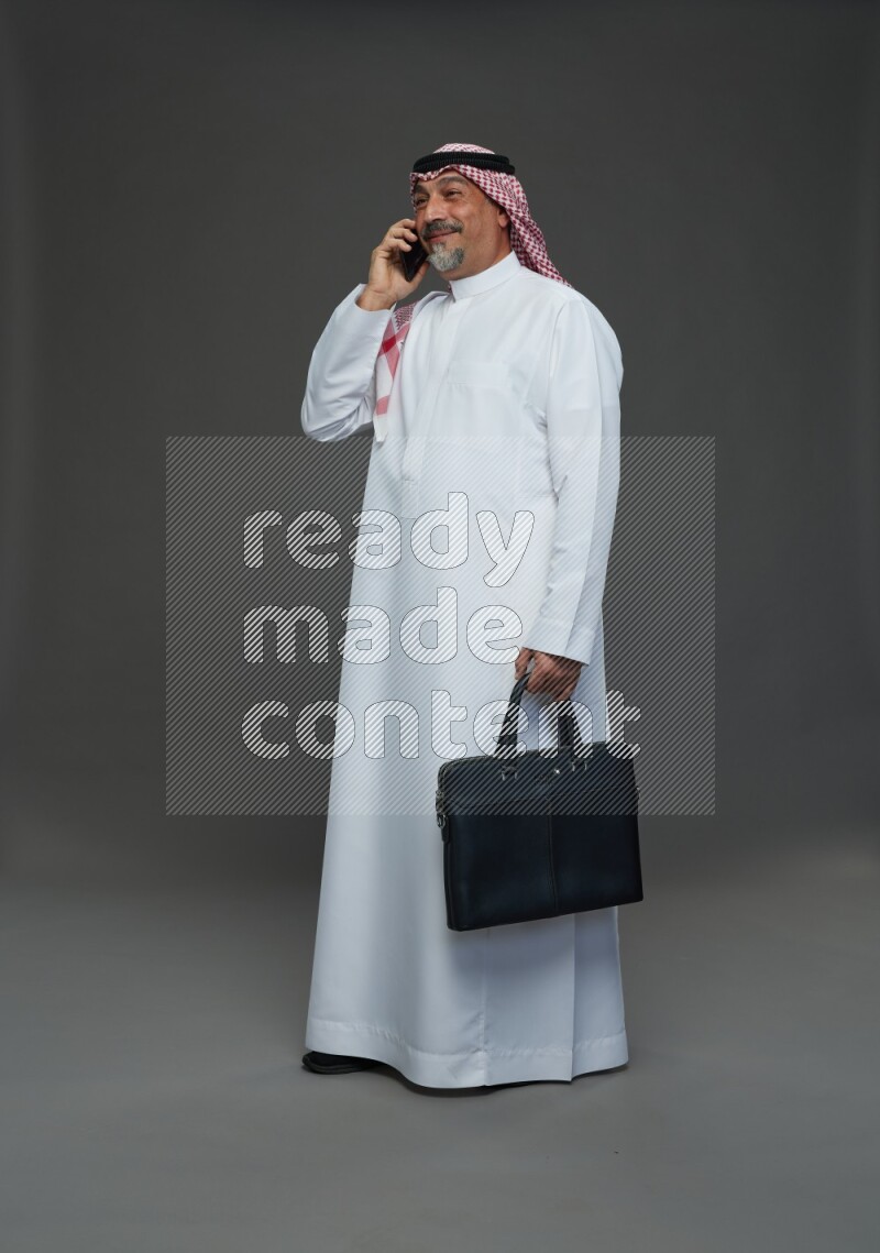 Saudi man with shomag Standing holding bag talking on phone on gray background