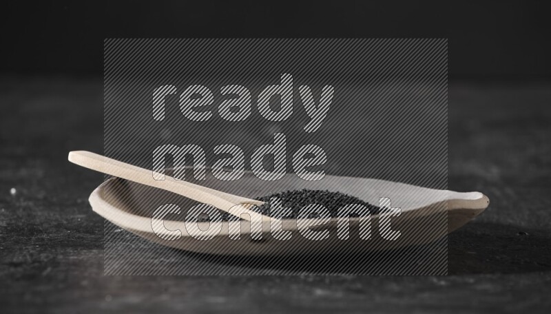 A multicolored pottery plate full of black seeds and wooden spoon full of seeds on a textured black flooring