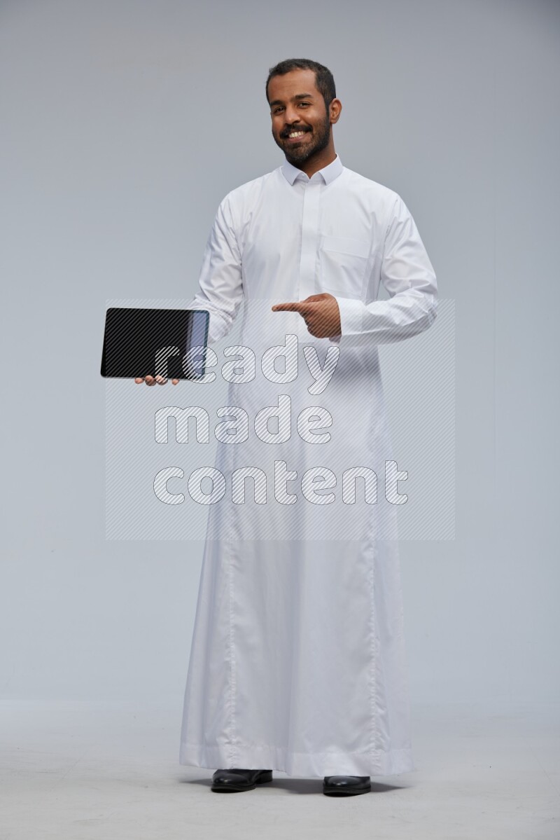Saudi man wearing Thob standing showing tablet to camera on Gray background