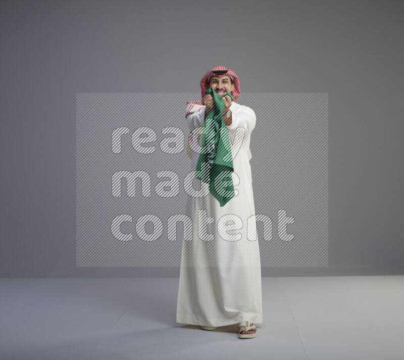 A saudi man standing wearing thob and red shomag holding big saudi flag on gray background