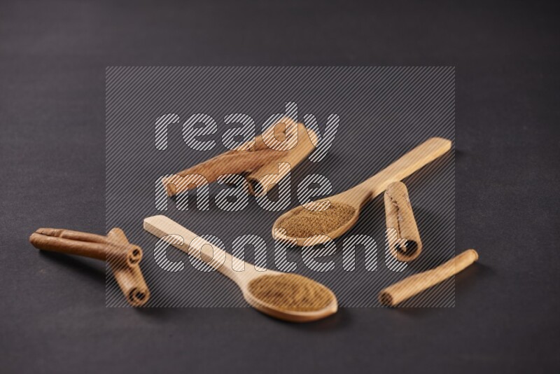 Two wooden spoons full of cinnamon powder with cinnamon sticks on black background