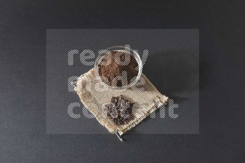 A glass bowl full of cloves powder with cloves grains on a burlap piece on a black flooring