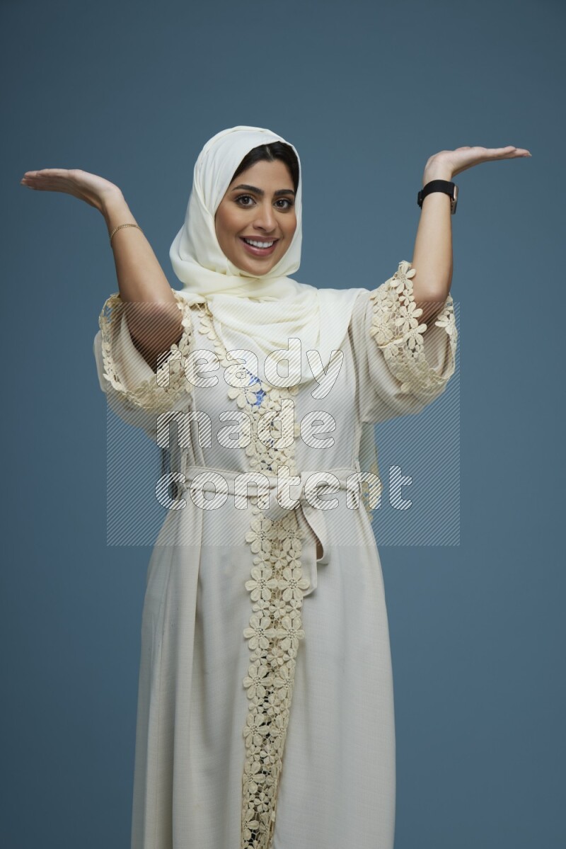 A Saudi woman pointing in a blue background wearing an off-white Abaya Hijab