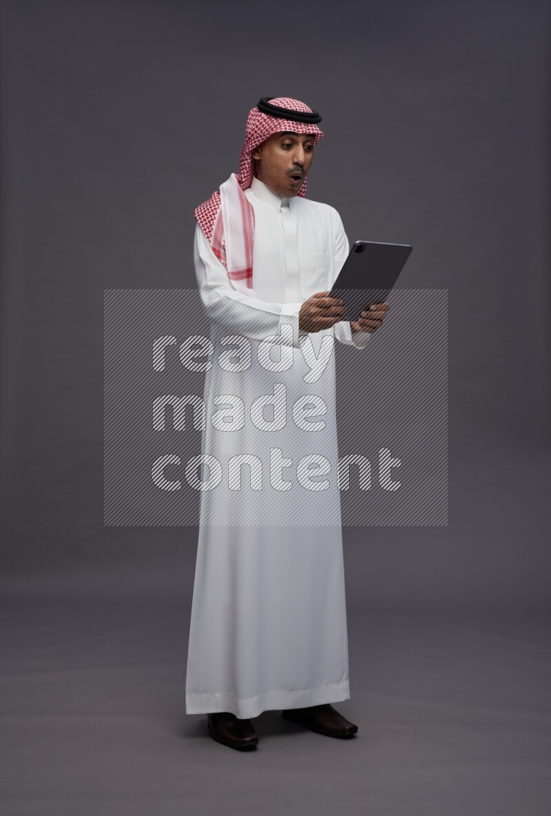 Saudi man wearing thob and shomag standing working on tablet on gray background