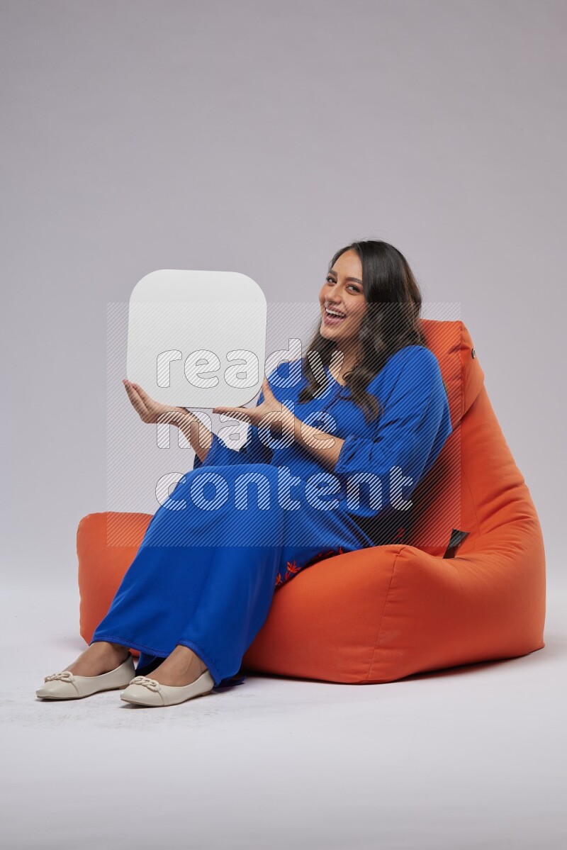 A Woman sitting on an orange beanbag wearing Jalabeya holding a social media sign
