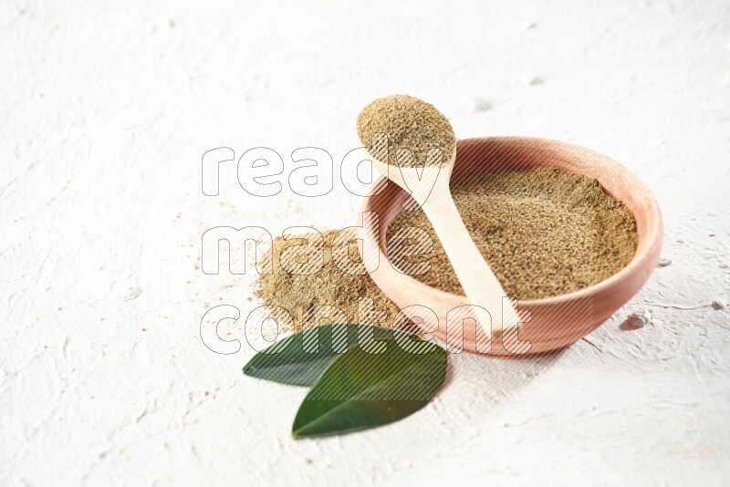 A wooden bowl and wooden spoon full of cumin powder on textured white flooring