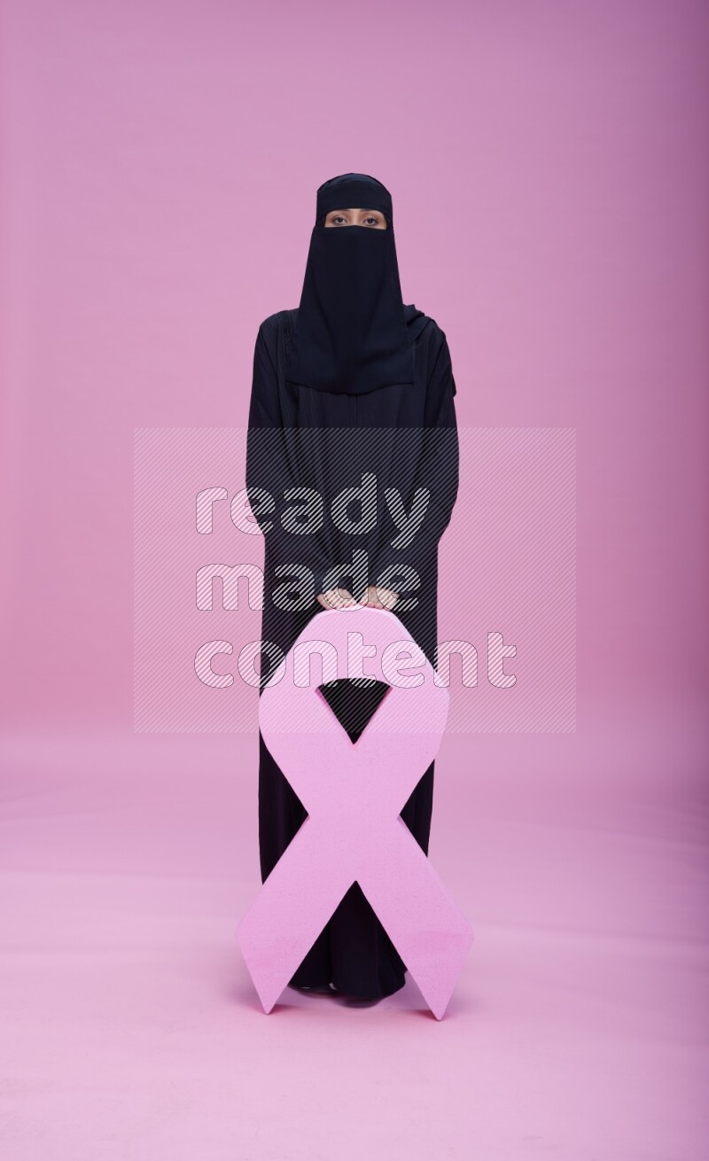 Saudi woman wearing abaya and niqab standing holding awareness ribbon on pink background