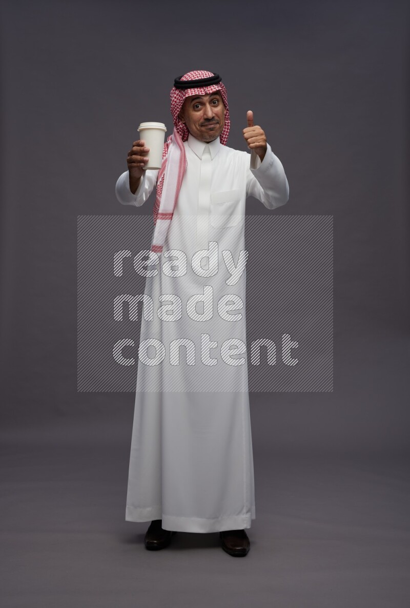 Saudi man wearing thob and shomag standing holding paper cup on gray background