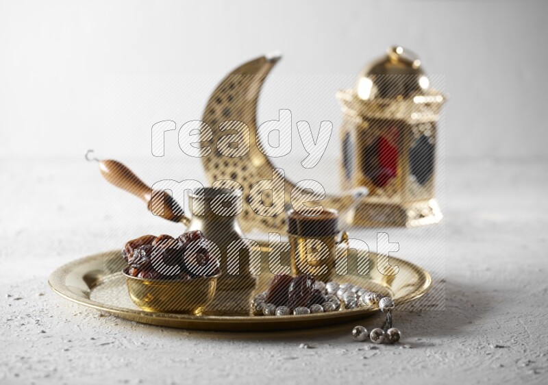 Dates in a metal bowl with coffee and prayer beads on a tray beside lanterns in a light setup