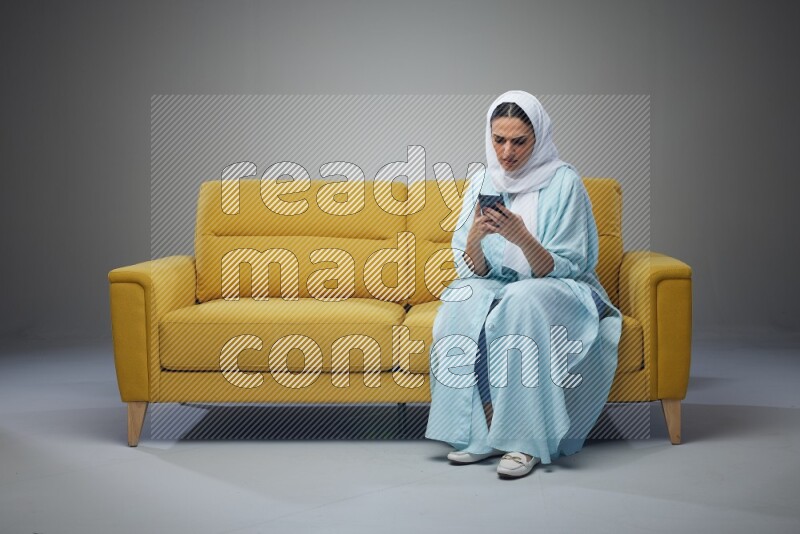 A Saudi woman wearing a light blue Abaya and white head scarf sitting on a yellow sofa and using her phone eye level on a grey background
