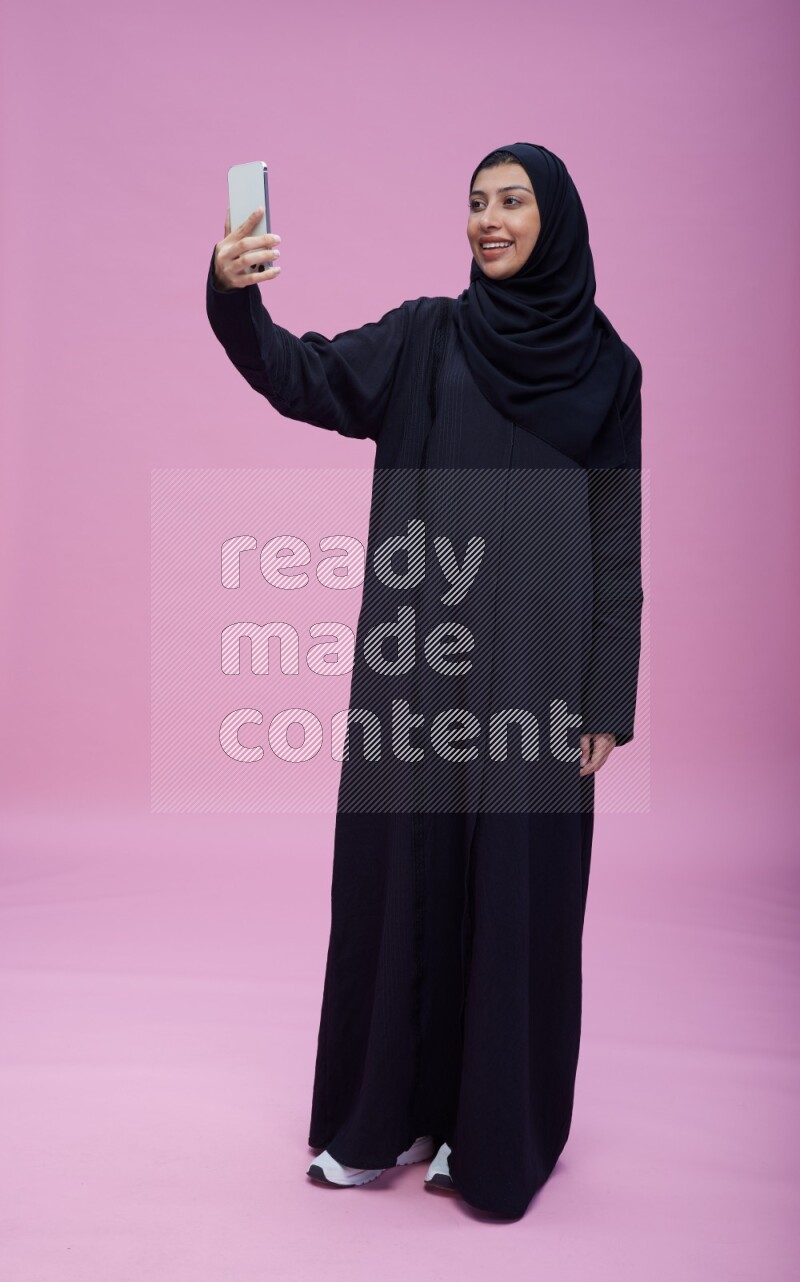 Saudi woman wearing Abaya standing taking selfie on pink background