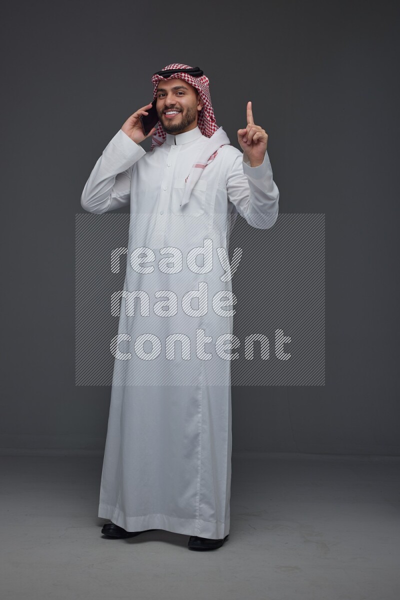 A Saudi man wearing Thobe and Shmagh talking in the phone eye level on a gray background